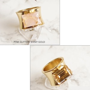 May include: Two rings with wide bands. The top ring is shiny gold with a rectangular pink glitter stone, labeled PINK GLITTER | SHINY GOLD. The bottom ring is matte gold with a rectangular amber-colored stone, labeled GOLDEN SHADOW | MATTE GOLD.