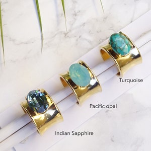 May include: Three gold-toned wide band rings, each featuring a large oval-shaped stone. The rings are labelled Indian Sapphire, Pacific Opal, and Turquoise, corresponding to the dark blue, light blue, and marbled teal stones set in the metal.