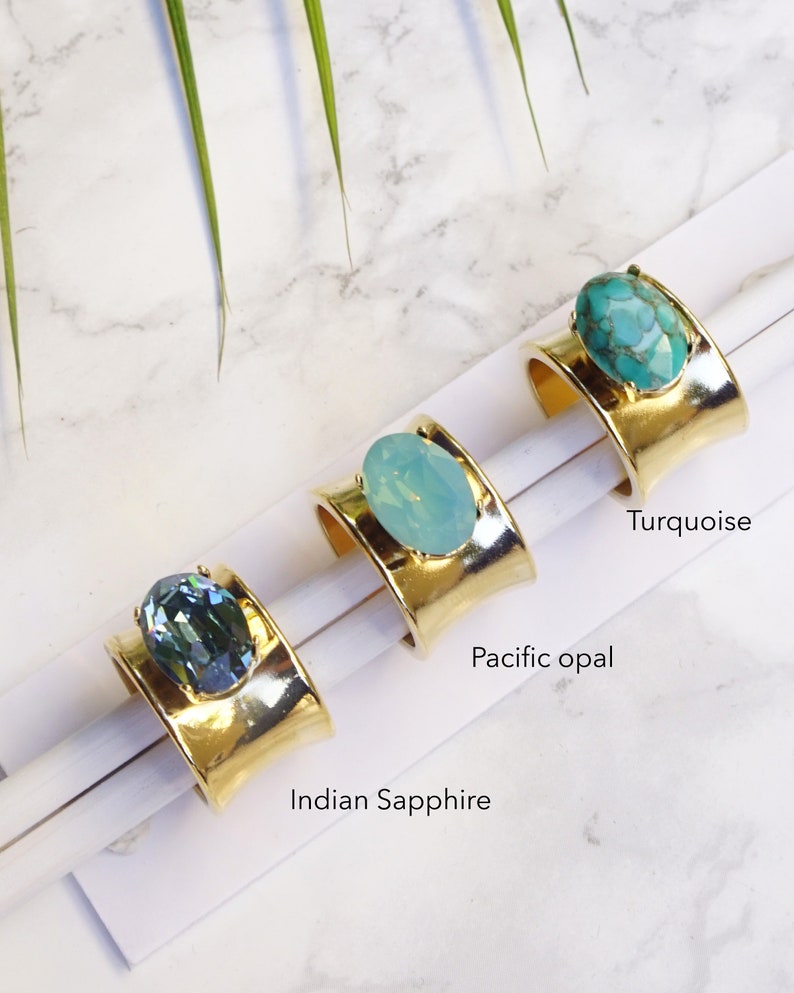 May include: Three gold-toned wide band rings, each featuring a large oval-shaped stone. The rings are labelled Indian Sapphire, Pacific Opal, and Turquoise, corresponding to the dark blue, light blue, and marbled teal stones set in the metal.