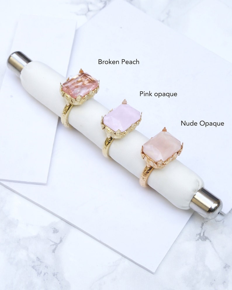 May include: Three gold rings with different colored gemstone settings. The rings are displayed on a white surface. The gemstones are labeled "Broken Peach", "Pink opaque", and "Nude Opaque".