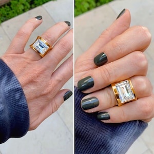 May include: Two hands wearing gold rings with large clear rectangular stones. The nails are painted a dark green color.
