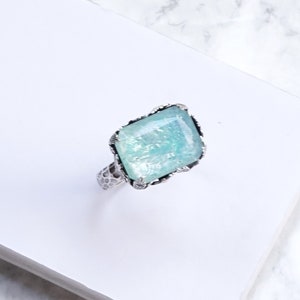 K&ouml;nnte beinhalten: Ein quadratischer Ring mit einem hellblauen Edelstein, der in einer antiken Silberfassung gefasst ist. Der Ring hat ein strukturiertes Band.
