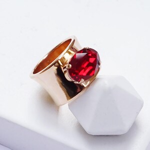 May include: A wide rose gold band ring featuring a large, oval-shaped, faceted deep red stone set in a prong setting. The ring is positioned on a white surface with the text SIAM | ROSE GOLD printed in black at the bottom right corner.