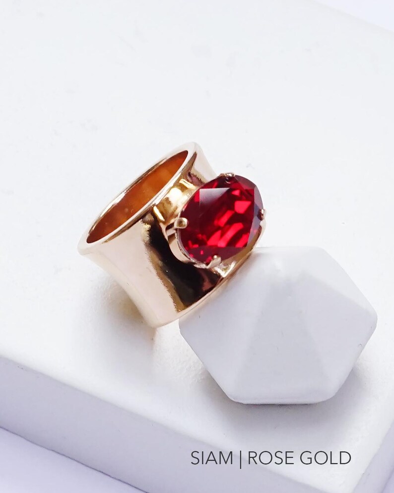 May include: A wide rose gold band ring featuring a large, oval-shaped, faceted deep red stone set in a prong setting. The ring is positioned on a white surface with the text SIAM | ROSE GOLD printed in black at the bottom right corner.