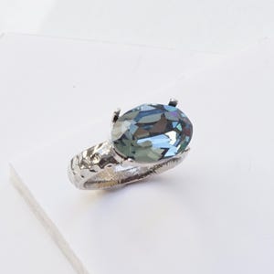 May include: A silver-toned ring featuring a large, oval-cut Indian Sapphire crystal set in a four-prong mount. The band has a textured, hammered finish. The text Indian Sapphire is printed in black at the bottom left of the image frame.