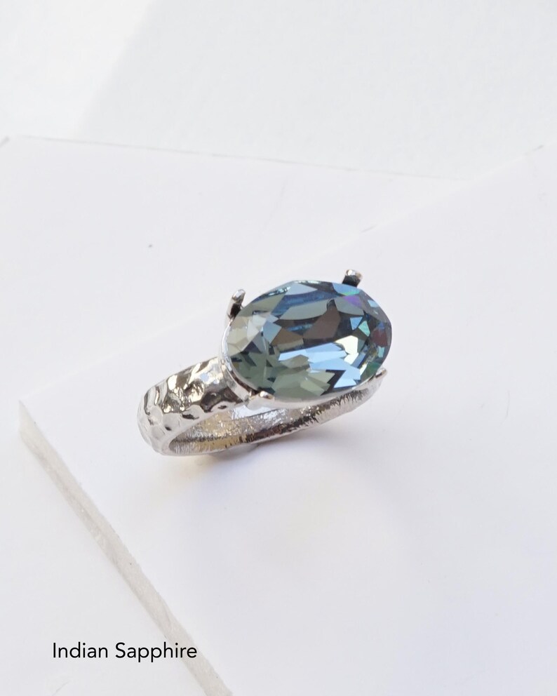 May include: A silver-toned ring featuring a large, oval-cut Indian Sapphire crystal set in a four-prong mount. The band has a textured, hammered finish. The text Indian Sapphire is printed in black at the bottom left of the image frame.