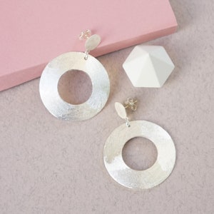 May include: A pair of silver earrings with a circular design. The earrings are made of white matte silver and have a textured finish.