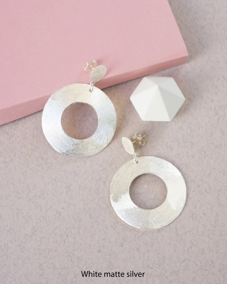 May include: A pair of silver earrings with a circular design. The earrings are made of white matte silver and have a textured finish.