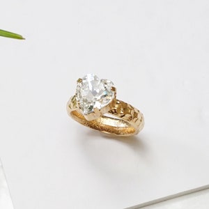 May include: A ring featuring a clear heart-shaped crystal set in a rose gold finish band with a textured, hammered surface. The band has a prong setting holding the stone securely. The text Clear crystal with Rose gold finish is visible.