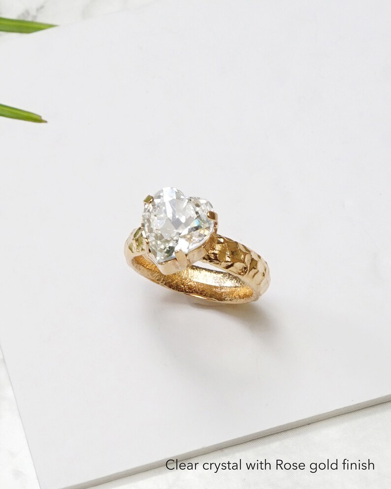 May include: A ring featuring a clear heart-shaped crystal set in a rose gold finish band with a textured, hammered surface. The band has a prong setting holding the stone securely. The text Clear crystal with Rose gold finish is visible.