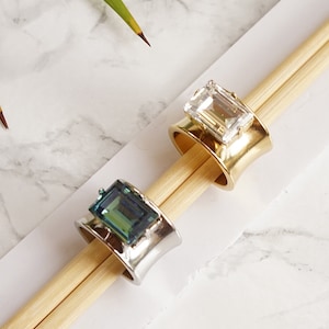 May include: Two rings with rectangular stones. One ring is shiny silver with an erinite green stone. The other ring is matte gold with a clear crystal stone. Text below reads: ERINITE SHINY SILVER and CLEAR CRYSTAL MATTE GOLD.