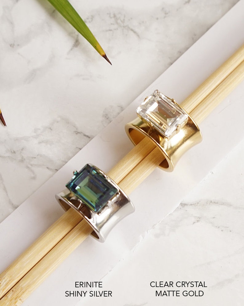 May include: Two rings with rectangular stones. One ring is shiny silver with an erinite green stone. The other ring is matte gold with a clear crystal stone. Text below reads: ERINITE SHINY SILVER and CLEAR CRYSTAL MATTE GOLD.