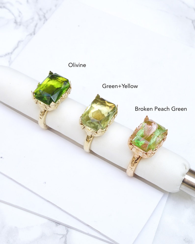 May include: Three gold rings with different colored green glass stones. The rings are labeled "Olivine", "Green+Yellow", and "Broken Peach Green".