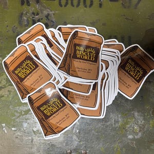 May include: A collection of stickers featuring a brown paper bag design with the text "Billy's Biscuit World" and the phrase "West by God Virginia." The stickers also include the location "Charleston, WV" and the date "April 22nd, 2024."