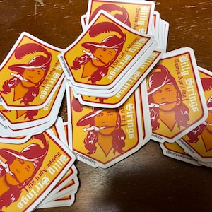 Puede incluir: Un montón de pegatinas con una ilustración estilizada de una persona con sombrero y espada, en tonos rojos, naranjas y amarillos. El texto "Billy Strings Tampa April 8th" está impreso en cada pegatina.