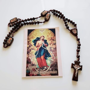 Peut inclure: Un chapelet marron foncé avec des perles ovales et triangulaires, un crucifix et une carte avec l'image d'une femme en robes bleues et rouges. La carte porte le texte "Novena Virgen Desatanudos".