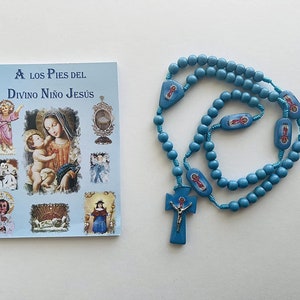May include: A light blue rosary with a cross pendant and a small booklet titled "A Los Pies Del Divino Niño Jesús". The rosary features blue beads and decorative rectangular pieces with religious imagery.
