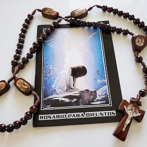 May include: A brown wooden rosary with a small wooden cross featuring a depiction of Jesus Christ. The rosary is accompanied by a small booklet with the text "ROSARIO PARA DIFUNTOS" on the cover.