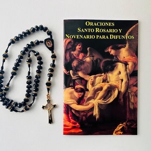 May include: Black rosary beads with a gold cross and a small image of the Virgin Mary. The rosary beads are next to a booklet titled "Oraciones Santo Rosario y Novenario Para Difuntos".