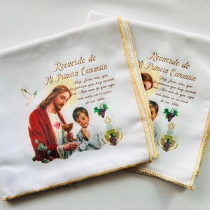 May include: Two white fabric napkins with gold trim and a religious image of Jesus giving communion to a young boy. The text on the napkins reads "Recuerdo de Mi Primera Comunión" which translates to "Memory of My First Communion".