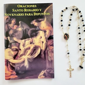 May include: A black and gold rosary with black beads and a gold cross. The rosary is next to a book titled "Oraciones Santo Rosario y Novenario Para Difuntos".