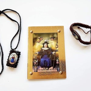 May include: A small, gold-colored booklet with a picture of a religious figure on the front. The booklet is surrounded by a black necklace with a pendant and a brown bracelet.