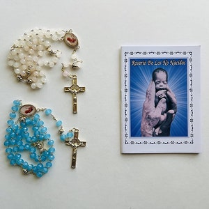 May include: Two rosaries, one with white beads and one with blue beads, with a gold cross and a small image of a baby. A booklet titled "Rosario De Los No Nacidos" is also included.