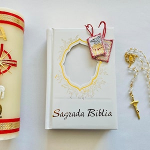 May include: A white candle with red and gold trim, a white book with gold lettering that says "Sagrada Biblia", and a white rosary with a gold cross.