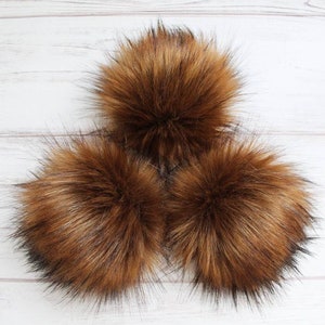 May include: Three brown faux fur pom poms on a white wooden background.
