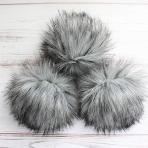 May include: Three fluffy gray pom poms on a white wooden background.