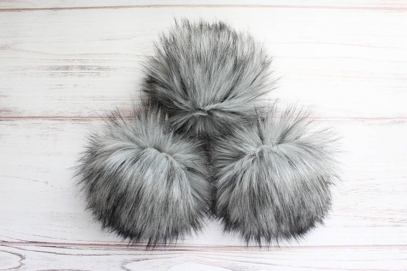 May include: Three fluffy gray pom poms on a white wooden background.