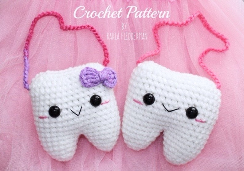 May include: Two white crocheted tooth-shaped plush toys with black eyes and smiling mouths. One tooth has a purple bow. Both are attached to a pink and purple crocheted strap.