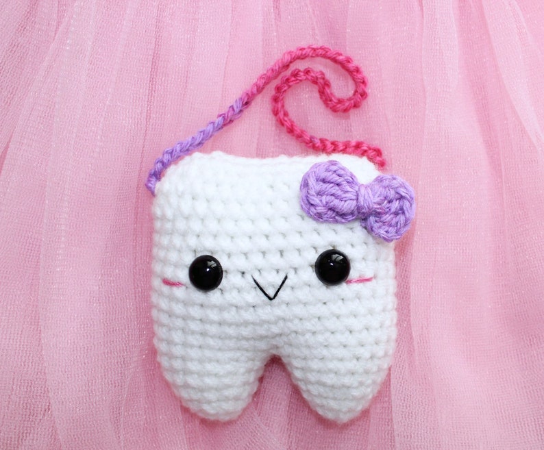 May include: White crocheted tooth with a purple bow, black eyes, and a pink and purple yarn strap.
