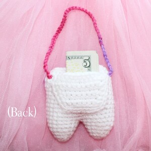 May include: A white crocheted tooth-shaped pouch with a pink and purple strap. A five dollar bill is inside the pouch. The pouch is on a pink tulle background.