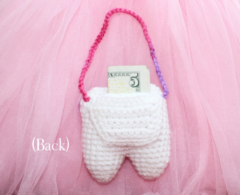 May include: A white crocheted tooth-shaped pouch with a pink and purple strap. A five dollar bill is inside the pouch. The pouch is on a pink tulle background.