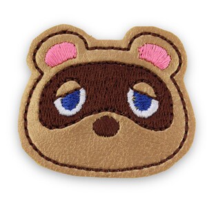 May include: A tan faux leather patch shaped like a raccoon head with embroidered details. The design features dark brown fur markings, pink inner ears, and blue and white eyes. The entire patch is outlined with dark brown stitching.