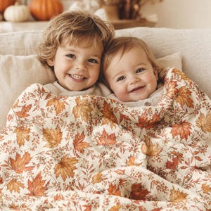 May include: A soft, cream-coloured throw blanket featuring a detailed autumnal pattern of orange, red, and brown maple leaves interspersed with small berry sprigs. The fabric has a textured appearance and is draped over two smiling children.