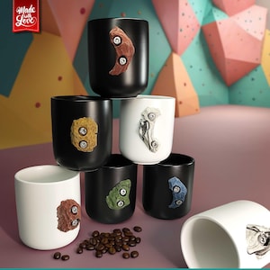 Könnte beinhalten: Eine Ansammlung schwarzer und weißer Keramikbecher, die jeweils mit einem Klettergriff-Design in verschiedenen Farben verziert sind. Die zylindrischen Becher stehen auf einer Oberfläche mit Kaffeebohnen. Das Bild enthält den Text "Made with Love".
