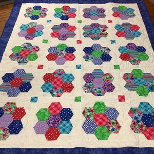 May include: A white quilt with a blue border. The quilt is made up of a repeating pattern of hexagonal flower shapes in shades of pink, blue, green, and white.
