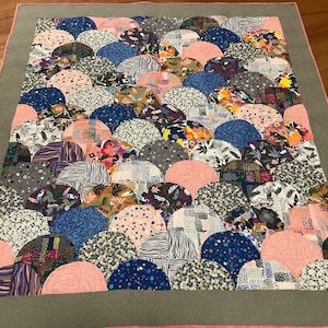 May include: A colorful quilt with a pattern of overlapping, semi-circular shapes in various shades of pink, blue, gray, and brown. The quilt is edged with a gray border and a pink trim.