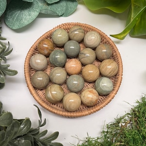 May include: A collection of 18 smooth, round, polished green and brown stones in a woven basket. The stones are arranged in a circular pattern.
