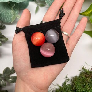 K&ouml;nnte beinhalten: Drei kleine, runde Edelsteinkugeln in einem schwarzen Samtbeutel. Die Kugeln sind rot, grau und rosa.