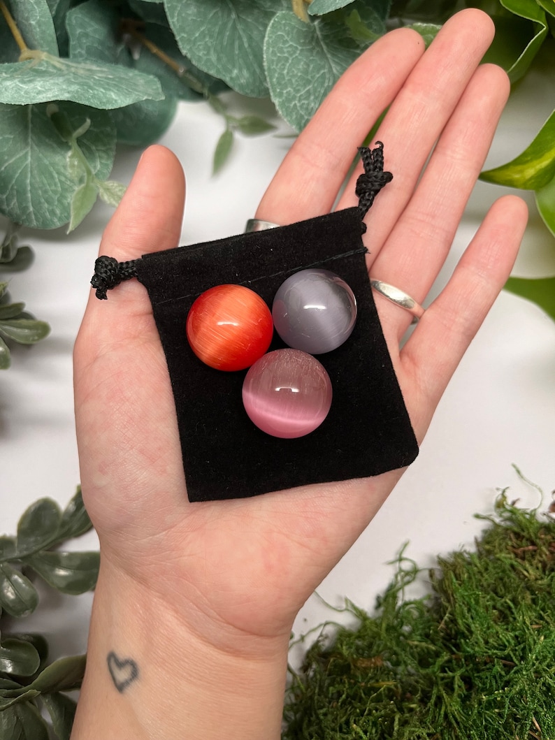 K&ouml;nnte beinhalten: Drei kleine, runde Edelsteinkugeln in einem schwarzen Samtbeutel. Die Kugeln sind rot, grau und rosa.