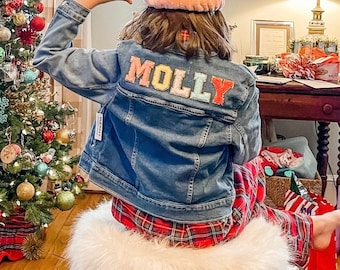 Könnte beinhalten: Ein Mädchen trägt eine blaue Jeansjacke mit dem Namen "MOLLY" in bunten Buchstaben auf dem Rücken. Sie trägt darunter ein rot-schwarzes kariertes Hemd und ein rosa Stirnband.
