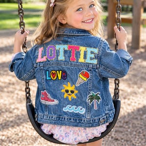 May include: A blue denim jacket featuring the name LOTTIE in large, colorful, glittery letters across the back. The jacket is decorated with various embroidered patches including the word LOVE, an ice cream cone, a sun, a sneaker, and waves.