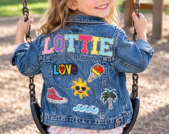May include: A blue denim jacket featuring the name LOTTIE in large, colourful, glittery letters across the back. The jacket is decorated with various embroidered patches including the word LOVE, an ice cream cone, a sun, a trainer, and waves.