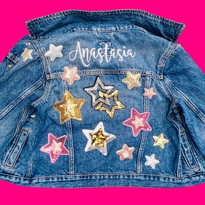 May include: A blue denim jacket with star-shaped patches in various colors, including gold, pink, and silver. The name "Anastasia" is written in cursive script across the upper back. The jacket has a classic collar and button closures.