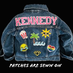 Peut inclure: Une veste en jean bleue avec divers patchs cousus sur le dos. Les patchs comprennent un soleil avec des lunettes de soleil, un palmier, une vague, un diamant, un seau de pop-corn, un cœur avec le texte "Love is Love" en couleurs arc-en-ciel et un emoji qui rit. Le texte "KENNEDY" est cousu au dos en lettres roses.