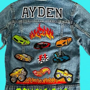 May include: A blue denim jacket with the name "AYDEN" sewn on the back. The jacket is decorated with various colorful patches, including cars, flames, and a checkered flag. The bottom of the jacket has the words "SEW IN ON" in green letters.