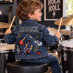 May include: A dark blue denim jacket featuring the word ROCK in white letters across the back, accompanied by embroidered patches of two electric guitars, a pair of headphones, musical notes, and a small red trumpet on the back panel.